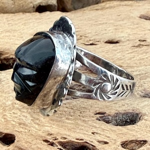 May include: A silver ring featuring a carved black stone face set in a scalloped bezel. The band is split into three sections, with the side displaying a circular sunburst pattern and etched arrow details along the metal surface of the ring.