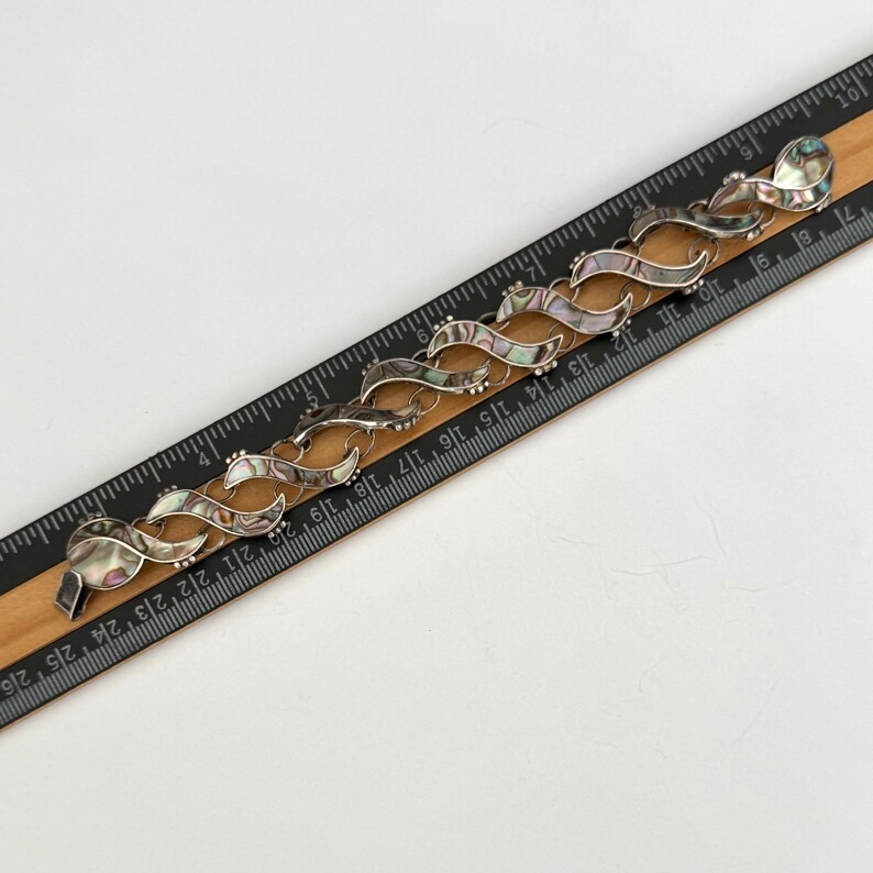 May include: A silver-toned bracelet with a wave-like design, featuring iridescent abalone shell inlays. The bracelet is shown on a wooden ruler, with measurements in inches.