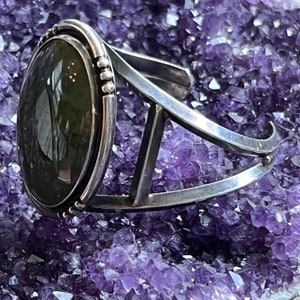 May include: A silver cuff bracelet featuring a large oval green gemstone set in a silver bezel. The bracelet is displayed on a bed of purple amethyst crystals.