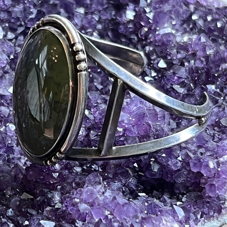 May include: A silver cuff bracelet featuring a large oval green gemstone set in a silver bezel. The bracelet is displayed on a bed of purple amethyst crystals.