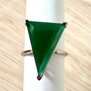 May include: A ring featuring a large, triangular, deep green gemstone set in a polished silver-toned metal band. The stone is oriented with the flat edge at the top and a pointed tip at the bottom, secured by small metal prongs at the corners.