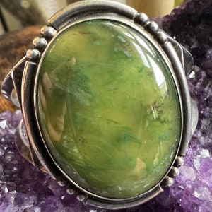 May include: A silver ring featuring a large, oval green stone set in the centre. The stone has a mottled, green and brown pattern.