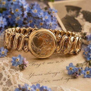 May include: A gold-tone metal expansion bracelet featuring a round central medallion with an engraved heart and floral motif. The band consists of interlocking textured links. The bracelet is displayed alongside blue forget-me-not flowers.
