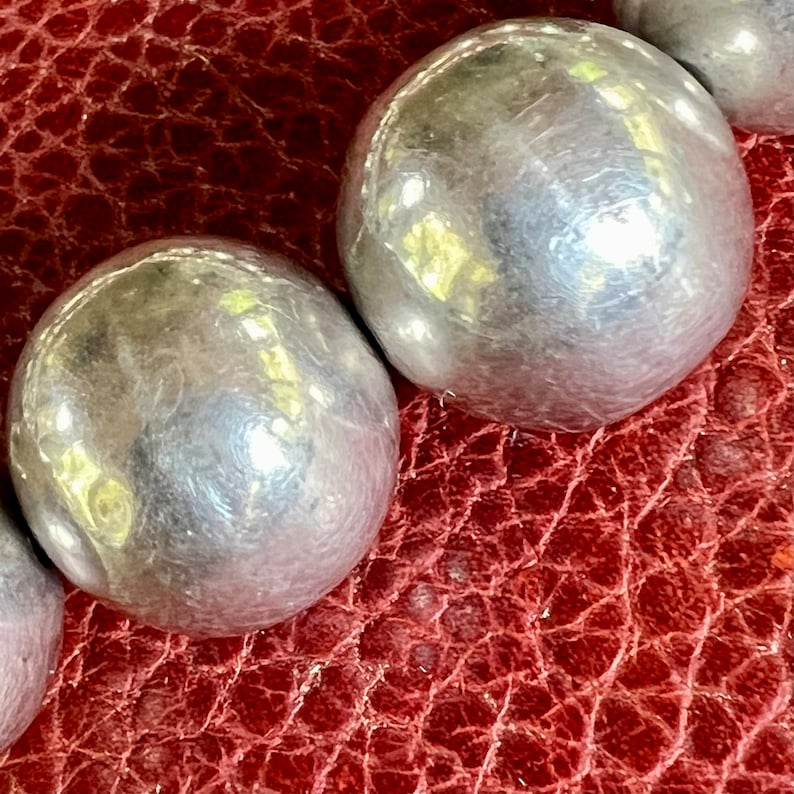 May include: A close-up of a silver beaded necklace with large, round beads. The beads are smooth and shiny, and they reflect the light.