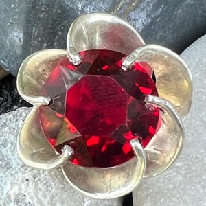 May include: A silver ring with a large, red, faceted gemstone set in a flower-shaped design.