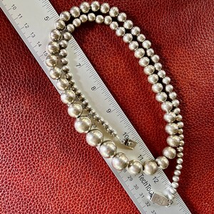 May include: A silver necklace with large round beads. The necklace is laying on a red leather surface with a ruler next to it.