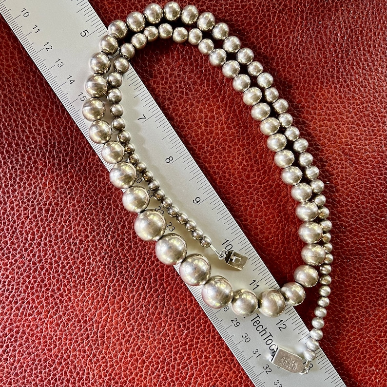 May include: A silver necklace with large round beads. The necklace is laying on a red leather surface with a ruler next to it.