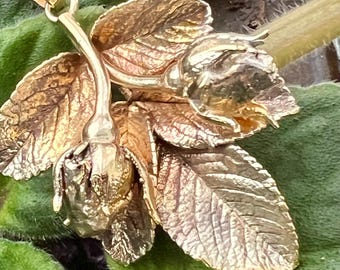 May include: A gold-toned pendant featuring a detailed floral design with two rose buds and several leaves. The pendant is suspended from a delicate chain, showcasing intricate textures and a naturalistic aesthetic. The background is a blurred green.