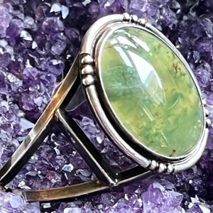 May include: A silver ring featuring a large oval green gemstone set in a bezel setting. The ring is displayed on a bed of purple amethyst crystals.