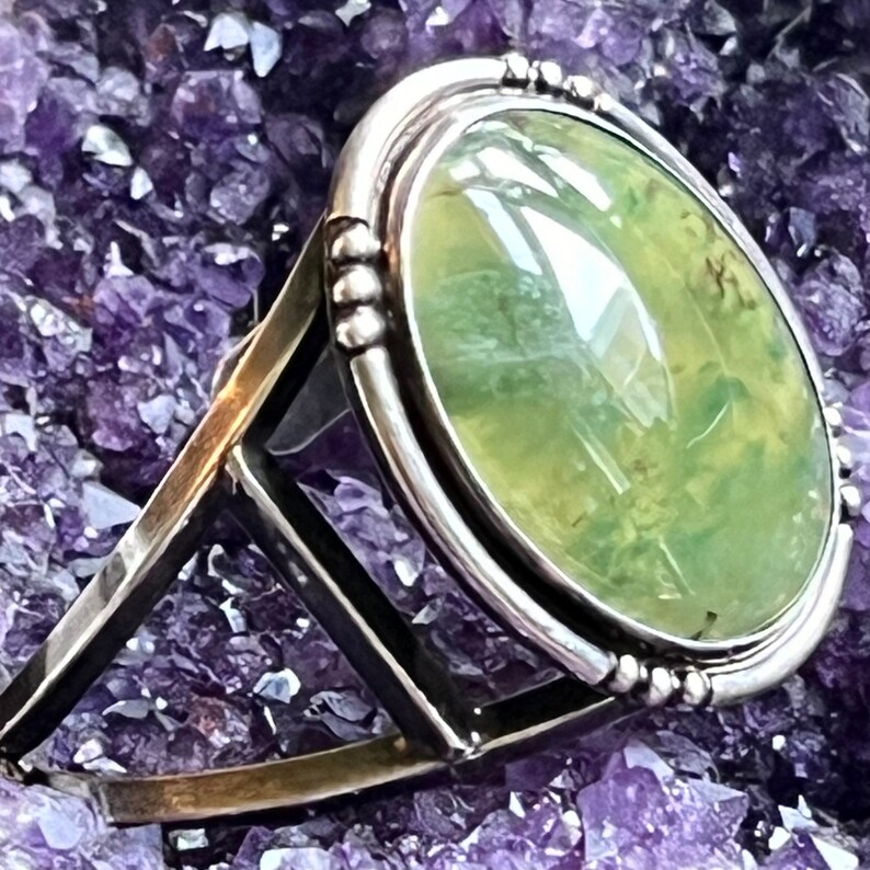 May include: A silver ring featuring a large oval green gemstone set in a bezel setting. The ring is displayed on a bed of purple amethyst crystals.