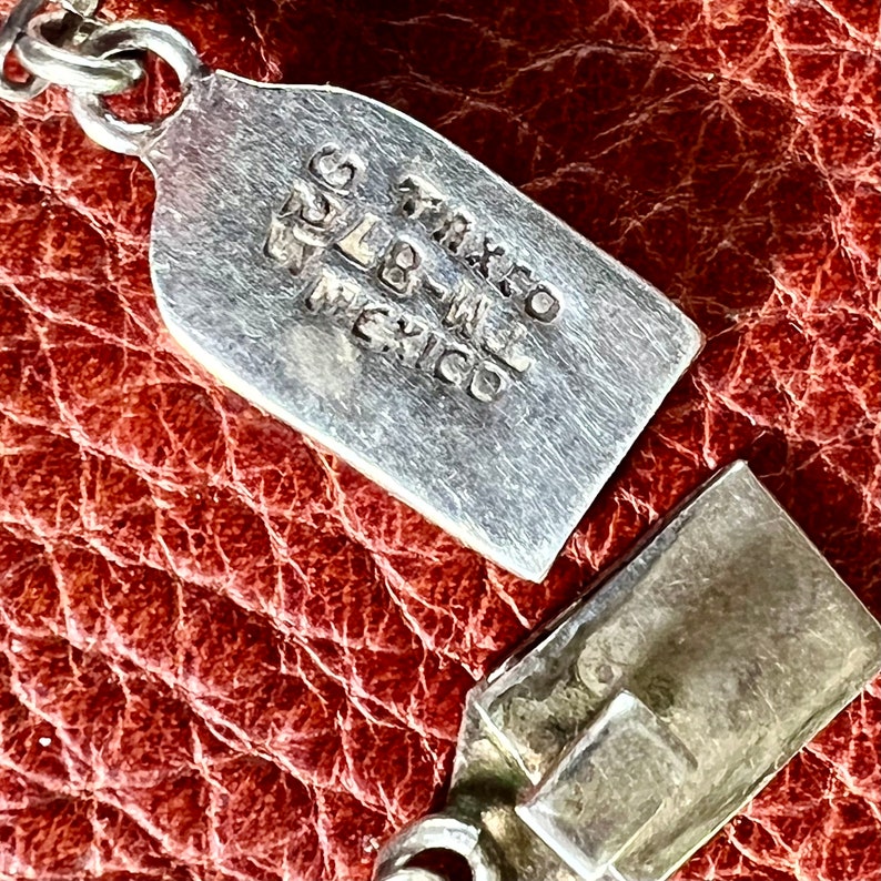 May include: Close-up of a silver charm with the text "TAXCO" "925" "MEXICO" stamped on it. The charm is on a brown leather surface.