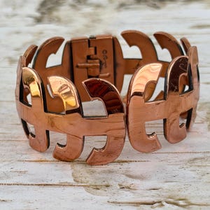 May include: A copper-toned cuff bracelet with an openwork design. The bracelet features a series of stylized letter "A" shapes, connected to form a hinged band. The clasp is visible, and the metal has a polished, reflective surface.