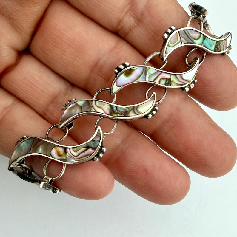 May include: A silver bracelet with iridescent abalone shell inlays. The bracelet features a series of curved, wave-like links, each adorned with the colorful shell. The links are connected by small, circular rings. The bracelet is displayed on a light background.