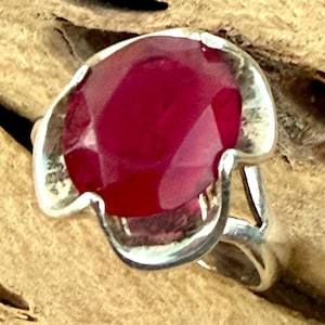 May include: A sterling silver ring featuring a large, oval-cut ruby gemstone. The ruby is a deep red color and is set in a bezel setting. The ring has a simple, elegant design.