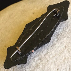 May include: Black enamel pin with a decorative design and a metal pin back.