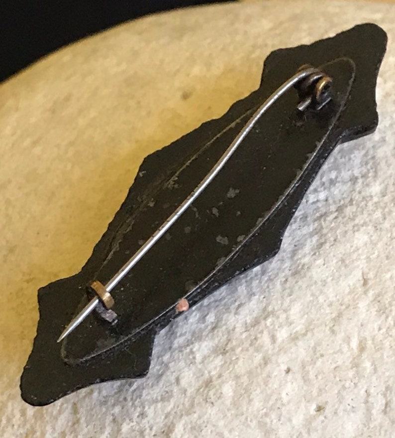 May include: Black enamel pin with a decorative design and a metal pin back.