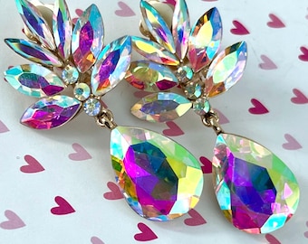 May include: A pair of iridescent crystal earrings. Each earring features a cluster of leaf-shaped crystals and a large, teardrop-shaped crystal pendant. The crystals reflect a rainbow of colors, set in gold-toned metal.