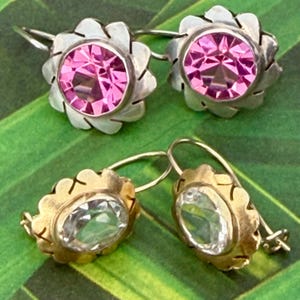 May include: Two pairs of earrings. The top pair features pink gemstones set in silver flower-shaped settings. The bottom pair has clear gemstones in gold-colored, scalloped settings. The earrings are displayed on a green surface.