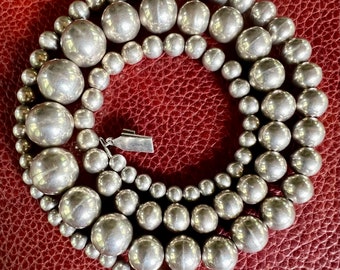 May include: A silver necklace with large, round beads. The necklace is made of multiple strands of beads, and the beads are all the same size. The necklace is lying on a red leather surface.