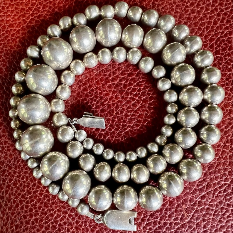 May include: A silver necklace with large, round beads. The necklace is made of multiple strands of beads, and the beads are all the same size. The necklace is lying on a red leather surface.