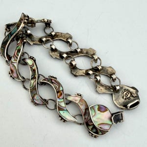 May include: A silver bracelet with iridescent abalone shell inlays. The bracelet features a series of curved, wave-like links connected by smaller chain links. The clasp is a hook-and-eye style, and the bracelet is laid out on a white surface.