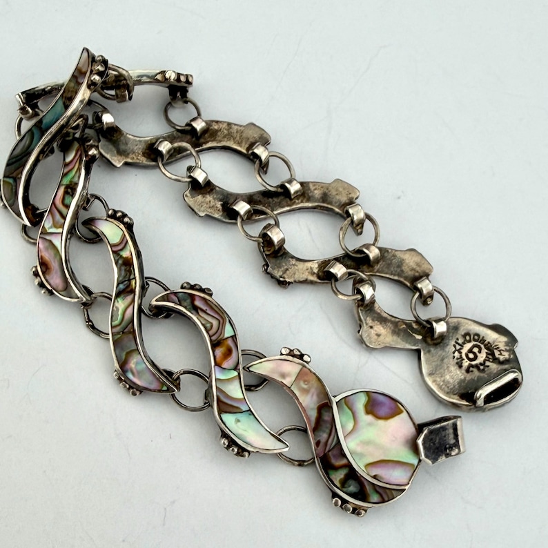 May include: A silver bracelet with iridescent abalone shell inlays. The bracelet features a series of curved, wave-like links connected by smaller chain links. The clasp is a hook-and-eye style, and the bracelet is laid out on a white surface.