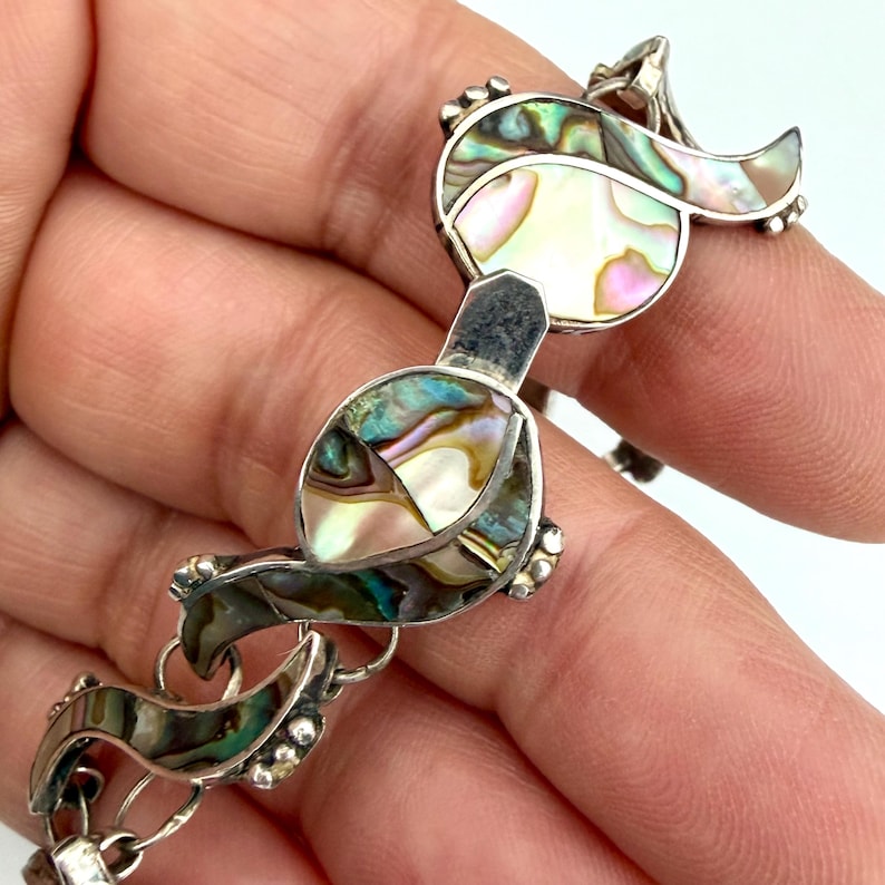 May include: A silver bracelet with iridescent abalone shell inlays. The bracelet features a series of stylized leaf-shaped links, each adorned with a mosaic of colorful shell fragments. The links are connected by small, round silver beads.