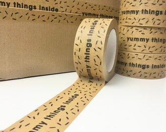 Puede incluir: Un rollo de cinta de embalaje marrón con un patrón repetitivo de guiones negros y el texto "Yummy things inside" impreso en él.