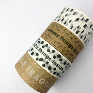 May include: Four rolls of washi tape with different designs. The top roll has a white background with black abstract shapes. The second roll has a brown background with white lines and the text "Yummy things inside". The third roll has a white background with black polka dots. The bottom roll has a brown background with white lines and the text "Handle with care".