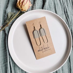 Könnte beinhalten: Ein Paar Ohrringe mit hellgrauem Lederaufsatz und goldfarbenem Metall-U-förmigem Unterteil. Die Ohrringe werden auf einer braunen Karte mit dem Text "white birch handmade goods" präsentiert. Die Ohrringe liegen auf einem weißen Teller, mit einer getrockneten Rose und einem hellblauen Tuch im Hintergrund.