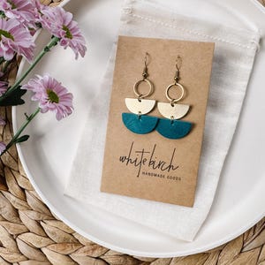 May include: Handmade earrings with a brass and teal design. The earrings are presented on a brown card with the text "white birch HANDMADE GOODS". The earrings are displayed on a white plate with flowers and a woven background.