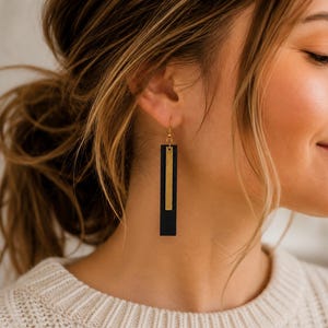 Peut inclure: Une boucle d'oreille pendante composée d'un long pendentif rectangulaire noir superposé à une barre métallique dorée plus fine et plus courte. Le bijou est suspendu à un crochet doré, formant un design géométrique vertical sur le lobe.