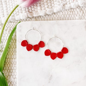 May include: A pair of silver hoop earrings with five bright red petal-shaped charms. The earrings are displayed on a white marble surface, with a pink tulip and white textured background.