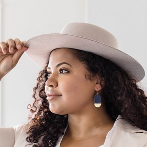 May include: A person wearing a light beige fedora hat and navy blue and gold earrings. The earrings feature a rectangular navy blue top and a gold half-circle bottom. The person is wearing a white shirt and a light beige blazer.