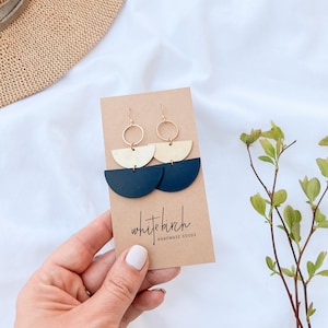May include: Gold and navy blue half-circle earrings with gold hoops, presented on a brown card with the text "white birch handmade goods". The earrings are held by a hand against a white backdrop.