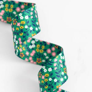 May include: A green ribbon with a floral pattern featuring small pink, yellow, and white flowers. The ribbon is curled, showcasing the detailed design of the flowers and leaves. The ribbon is likely made of fabric.