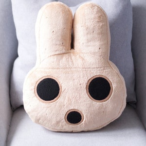 Op de afbeelding: Een pluche beige konijnenkussen met zwarte geborduurde ogen en een neus. Het kussen ligt op een lichtgrijze bank.