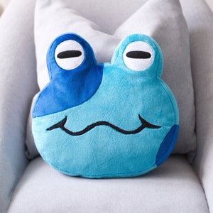 May include: A blue plush frog pillow with a smiling face. The pillow is sitting on a gray couch.