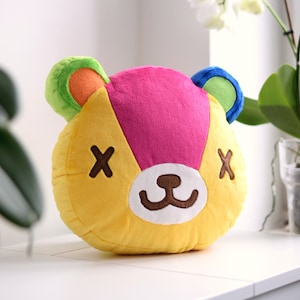 May include: A plush toy in the shape of a bear's head. The bear has a pink, yellow, green, and blue patchwork design. The bear has a white nose and brown eyes. The bear has an 'X' on each eye.
