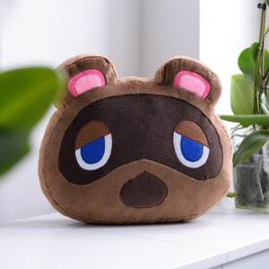 Op de afbeelding: Een pluche kussen in de vorm van Tom Nook uit Animal Crossing. Het kussen is bruin met een donkerbruin masker, roze binnenoren en blauwe ogen. Het kussen ligt op een wit oppervlak.