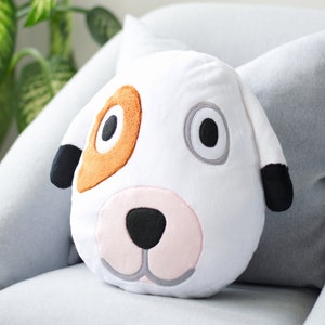 Op de afbeelding: Een wit pluche kussen in de vorm van een hondenkop met bruine, grijze en zwarte geborduurde details. Het kussen heeft grote zwarte ogen, een roze neus en een grijze mond.