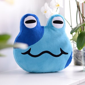 May include: A blue and light blue plush frog with a simple black smile. The frog has white eyes with black pupils.