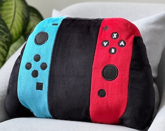 May include: A blue and red plush pillow shaped like a Nintendo Switch controller. The pillow has a black center section and features the buttons and joysticks of the controller.