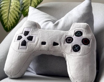May include: A grey plush pillow shaped like a video game controller. The pillow has black buttons and a grey background.