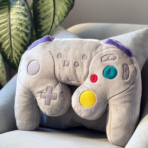 Può includere: Un cuscino di peluche grigio a forma di controller per videogiochi. Il controller ha accenti viola e pulsanti colorati.