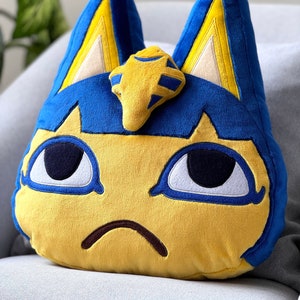 Op de afbeelding: Een pluche speelgoed van een blauwe en gele kat met een droevige uitdrukking. De kat heeft grote blauwe ogen en een gele neus. De kat draagt een gouden kroon.