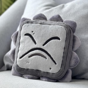 Op de afbeelding: Grijs pluche speelgoed in de vorm van een vierkant met een fronsend gezicht. Het speelgoed heeft een textuur en zit op een grijze bank.