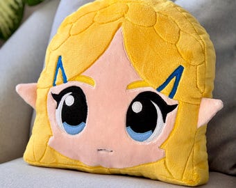 May include: A yellow plush pillow with a cartoon face of a girl with large eyes and blonde hair. The pillow is on a gray couch.