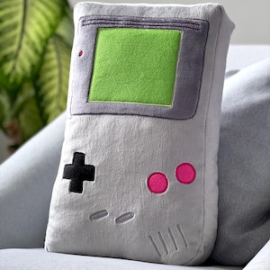 Puede incluir: Una almohada de felpa gris con forma de consola de videojuegos portátil. La almohada tiene una pantalla verde, botones negros y detalles rosas.