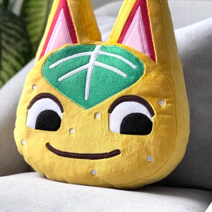 Op de afbeelding: Een geel pluche speelgoed in de vorm van een kat met een groen blad op zijn voorhoofd. De kat heeft zwarte ogen, een bruine glimlach en roze oren met rode accenten.